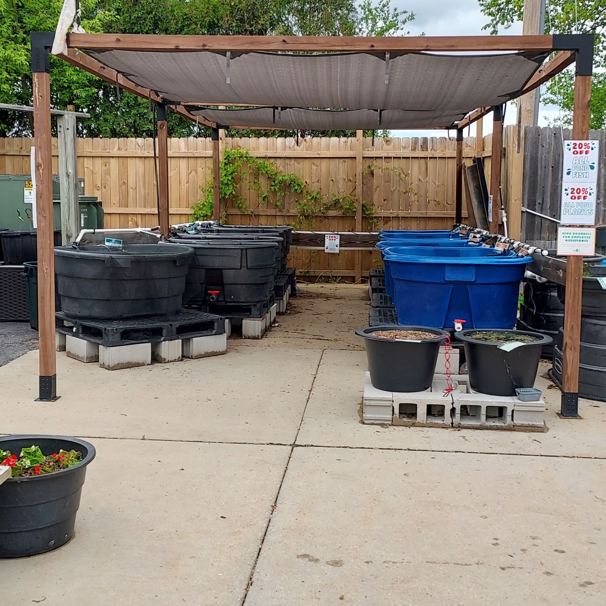 A covered outdoor area with large black and blue plastic tanks on concrete blocks, surrounded by a wooden fence. Small planters with plants are in the foreground, and signs are posted on the structures posts.