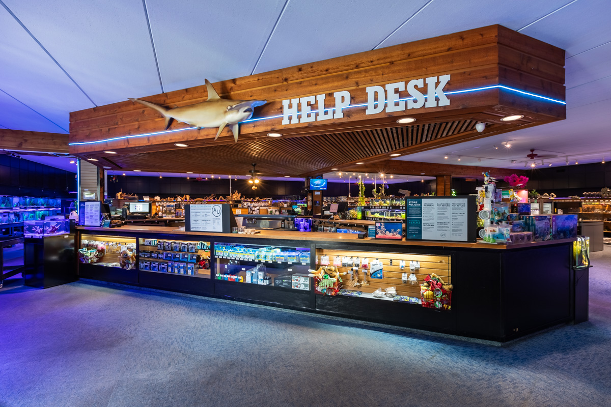The image shows an aquarium help desk with a wooden overhang, a shark model above, and bright lights. The counter displays brochures and souvenirs, with gift shop shelves and aquatic-themed items visible in the background.