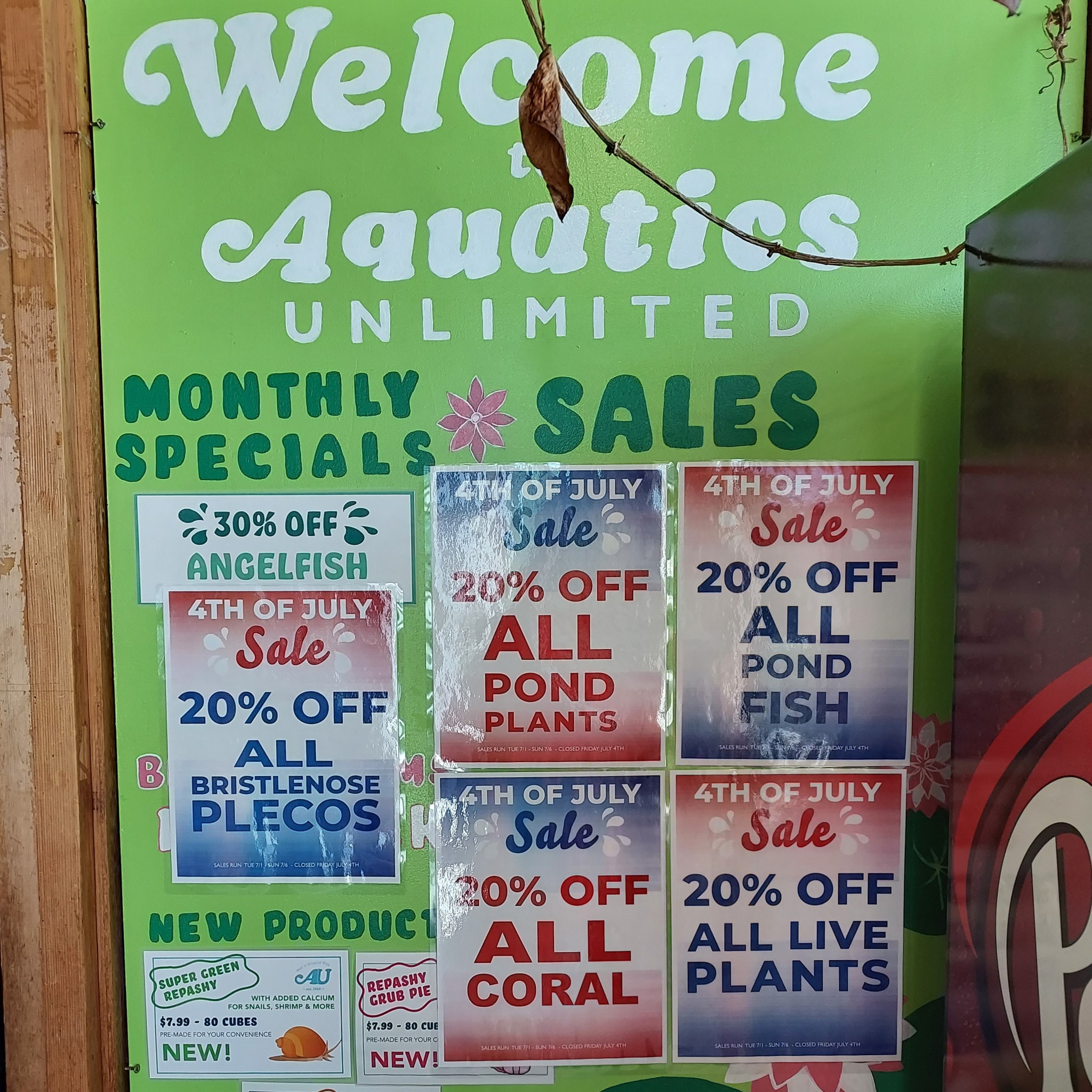 A green sign for Aquatics Unlimited shows monthly specials and 4th of July sales, including 20% off pond plants, pond fish, live plants, coral, bristlenose plecos, and 30% off angelfish.