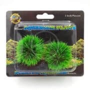 Two artificial green aquarium plants with spiky leaves are packaged in a clear plastic case labeled “3 Inch/Pouces” and “Aquarium Plant.” The background shows rocks and aquatic scenery.