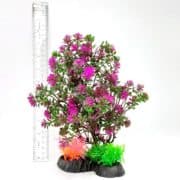 A plastic aquarium plant with pink flowers and green leaves stands next to a clear ruler for scale. The plant is anchored to a black base with small orange and green decorations at the bottom.