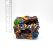 A small decorative treasure chest ornament with painted coins, gems, and a yellow eel, shown beside a ruler for scale against a white background. The chest appears weathered and has holes on its sides.