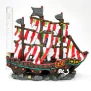 A colorful model pirate ship with red and white striped sails sits next to a vertical ruler for scale. The ship has three masts, gold accents, and rests on a rocky base. The background is plain white.