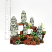 Five small stone statues with glasses and hats stand on brown brick-like steps with green plants around them. A clear ruler beside the display shows the height for scale. The background is white.