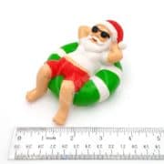 A small figurine of Santa Claus in sunglasses and red shorts relaxing on a green and white float, next to a ruler showing he is about 2 inches long.