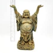 A small, weathered statue of a laughing Buddha with arms raised, standing on a decorative base. A ruler beside the statue shows it is about 7 inches tall. The background is plain white.