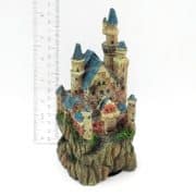 A detailed miniature castle with blue roofs sits atop a rocky base, with a ruler beside it showing the height is about 14 cm. The castle features towers, windows, and greenery accents.