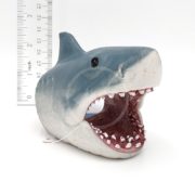 A small, open-mouthed shark figurine with visible teeth sits beside a ruler, indicating a height of about 2 inches. The model is painted gray on top and white on the bottom, with a black eye.