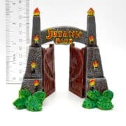 A small model of the Jurassic Park gate with open wooden doors, gray stone pillars topped with flames, green plants at the base, and a ruler beside it showing a height of about 4 inches.
