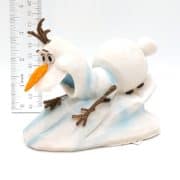 A small figurine of Olaf, the snowman from Frozen, sliding on his belly down a snowy slope. A ruler placed vertically behind shows Olaf is approximately 8 cm tall. The background is plain white.