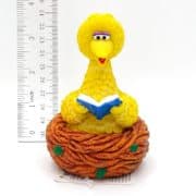 A small figurine of Big Bird from Sesame Street sits in a brown nest, reading a blue book. A ruler beside it shows the figure is about 3 inches tall.