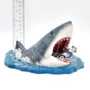 A small shark figurine with an open mouth holds a scuba tank in its jaws, resting on a blue water base. A ruler behind the figurine shows its height for scale.