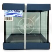 The SEAPORA RIMLESS CUBE TANK is a small glass aquarium housed in a dark blue cardboard frame with a label and barcode on the top left. The front glass displays a subtle white logo of two fish and waves.