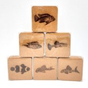 The DECORATIVE AQUARIUM FISH / LOGO COASTER SET 6 PCS includes six cork coasters stacked in a pyramid, each featuring a unique black illustration of an aquarium fish with diverse shapes and patterns on a simple white background, highlighting the intricate designs.