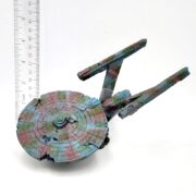 A weathered model spaceship, the UT Resin Sunken Space Ship LG Enterprise, sits next to a ruler for scale. Resembling underwater treasures, it features a multicolored patchy paint job with a classic sci-fi design, circular front, and elongated rear sections.
