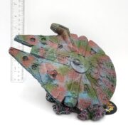 The UT RESIN SPACESHIP WRECK FALCON, a vibrant model evoking an underwater treasure, features pink, blue, and green hues in a textured pattern. It sits on a rocky base with a ruler in the background for scale.