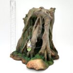 The UT RESIN MANGROVE CAVE is a realistic tree stump aquarium decor, crafted from durable resin with twisted roots and moss accents mimicking a mangrove cave. Its brown with green highlights, includes a small tag, and is displayed beside a ruler for height reference.