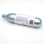 A metallic CO2 cartridge with a threaded nozzle lies on a white surface. A white label with text and symbols, including a barcode and recycling symbol, is attached to the side of the cartridge.