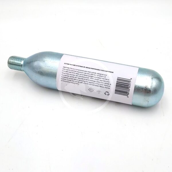 A metallic CO2 cartridge with a threaded nozzle lies on a white surface. A white label with text and symbols, including a barcode and recycling symbol, is attached to the side of the cartridge.