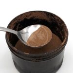 A metal spoon holds a scoop of fine brown powder above a black plastic container filled with the same powder, against a white background.