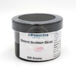 A small black plastic container labeled Aquatics Unlimited Brine Shrimp Eggs, 100 Grams sits on a white surface. The label includes a faint image of a shrimp and some text with instructions and ingredients.