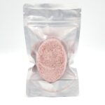 A pink oval sponge is sealed inside a clear, resealable plastic pouch standing upright against a white background.