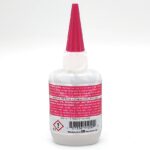A white bottle with a pink pointed cap, featuring warning labels and instructions in red and black text. The label includes hazard symbols and a barcode at the bottom. The product appears to be an adhesive or glue.