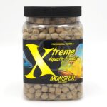 A clear plastic jar filled with round fish food pellets, labeled Xtreme Aquatic Foods Monster 9mm Pellet with images of two colorful fish and a large yellow X on the black label.