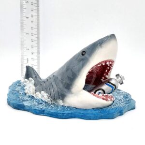 The PP RESIN JAWS WITH AIR TANK JWSR12 figurine features a shark bursting from blue water with an open mouth biting a scuba tank, with a ruler behind it to show scale. This piece is perfect for collectors seeking detailed, realistic designs.