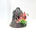 A cracked, weathered PP RESIN SUNKEN GARDENS BELL, adorned with vibrant artificial flowers and green vines, sits on a white surface with a clear ruler standing upright behind it for scale.