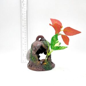 The PP RESIN SUNKEN GARDENS BELL is a small, brown, rock-like aquarium ornament with a hole, green moss accents, and red and green artificial plant leaves. A clear ruler stands upright behind it for scale.