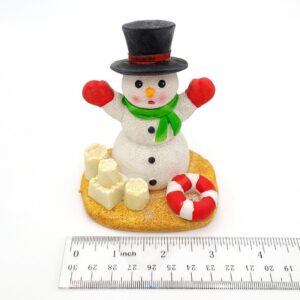 The PP RESIN HOLIDAY DECOR HOLR5B is a small, charming snowman figurine with a black hat, green scarf, and red mittens. It stands about 2 inches tall on sand beside sandcastles and a lifebuoy—perfect for festive decor.