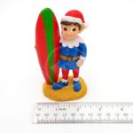 The PP RESIN HOLIDAY DECOR HOLR5B is a 3-inch tall Penn Plax figurine featuring a smiling elf in a blue and red Santa outfit standing by a festive surfboard on sandy ground.