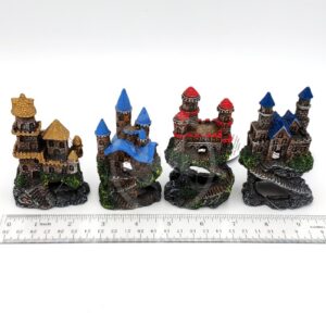 Four PP RESIN MINI CASTLE ASST RRW5CA decorations, each with colorful roofs (yellow, blue, red), are lined up before a ruler showing their 10–12 cm height. These resin castles feature arched doorways and detailed stonework.