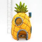 The PP RESIN SPONGEBOB PINEAPPLE 8 HOME W/ HOLES SBR41, with green leaves and blue-rimmed windows, sits next to a ruler for scale. It closely resembles SpongeBob SquarePants’ famous pineapple house.