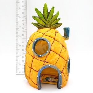 The PP RESIN SPONGEBOB PINEAPPLE 8 HOME W/ HOLES SBR41, with green leaves and blue-rimmed windows, sits next to a ruler for scale. It closely resembles SpongeBob SquarePants’ famous pineapple house.