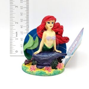 The PP RESIN ARIEL STRETCHING ON ROCK MEDIUM LMR6 is a colorful, detailed resin figurine of Ariel with red hair, green tail, and purple seashell top stretching on a rock. A ruler and card behind her display its medium size.