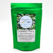A shiny green pouch labeled Complete Envy Balanced Shrimp Food by Shrimp Envy, featuring a circular logo with shrimp images and text describing it as a wholesome staple food for aquarium shrimp.